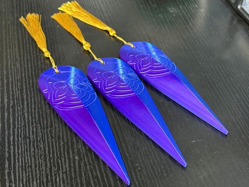 May include: Three gradient blue and purple bookmarks with a floral design and gold tassels. The bookmarks are elongated with a pointed end. The floral design is at the top of the bookmark. The tassels are attached to the top of each bookmark.
