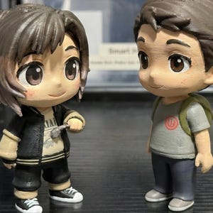 May include: Two anime-style figurines of young people. One wears a black jacket, black pants, and white sneakers, holding a small object. The other wears a gray t-shirt, blue pants, and gray shoes, with a green backpack. Both have brown hair and large eyes.