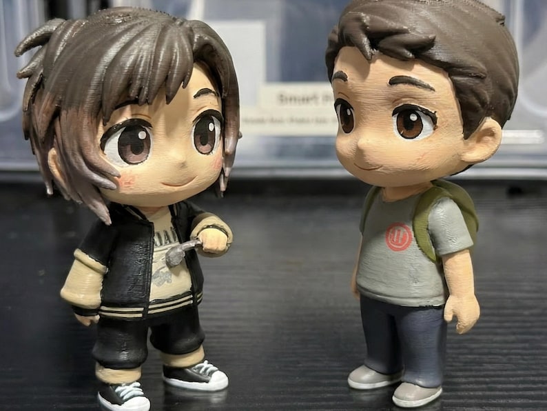 May include: Two anime-style figurines of young people. One wears a black jacket, black pants, and white sneakers, holding a small object. The other wears a gray t-shirt, blue pants, and gray shoes, with a green backpack. Both have brown hair and large eyes.