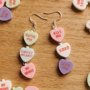 May include: Silver dangle earrings featuring heart-shaped candy charms with phrases like "Cutie Pie," "Sweet Heart," and "Be Mine." The earrings are set against a wooden background, surrounded by scattered candy hearts.