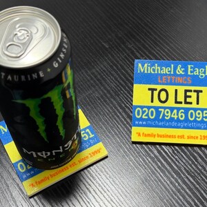 May include: A black and green Monster Energy drink can rests on a yellow and blue coaster. A second coaster with the text "TO LET" and a phone number is also visible. The coasters are square.
