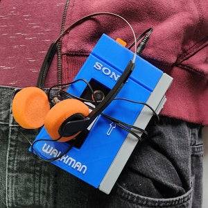 May include: A blue Sony Walkman cassette player with orange headphones. The Walkman is in a person's pocket.