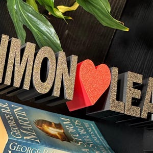 May include: Black wooden letters spelling "Simon" and "Leah" with gold glitter, separated by a red heart. The letters are on a shelf above a book.