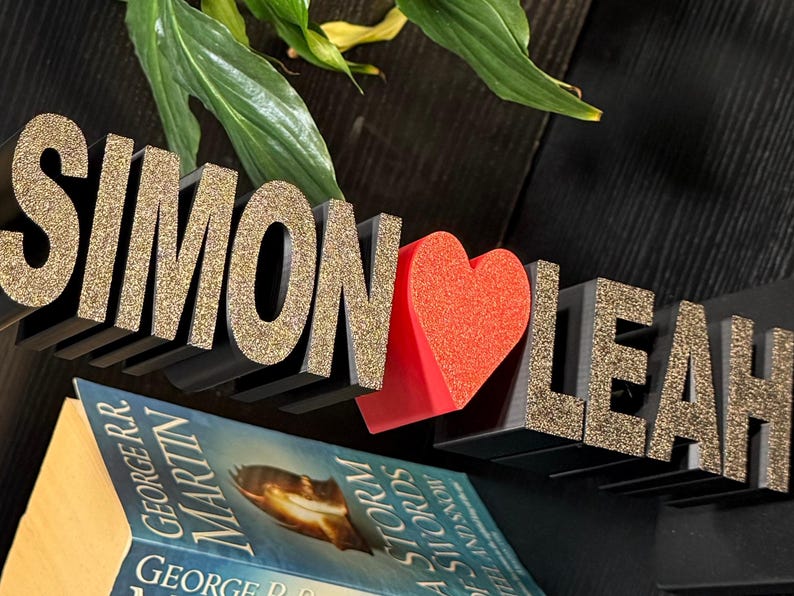 May include: Black wooden letters spelling "Simon" and "Leah" with gold glitter, separated by a red heart. The letters are on a shelf above a book.