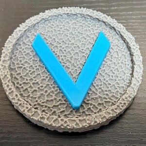 Puede incluir: Un objeto redondo gris con una superficie texturizada y una forma de "V" azul brillante en el centro. El objeto tiene un borde detallado, similar al encaje. El dise&ntilde;o es simple y moderno.