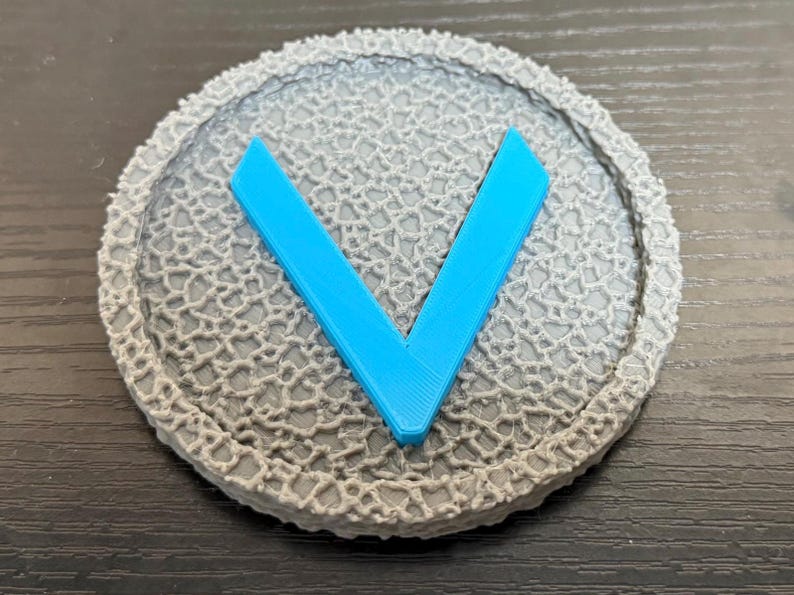 Puede incluir: Un objeto redondo gris con una superficie texturizada y una forma de "V" azul brillante en el centro. El objeto tiene un borde detallado, similar al encaje. El dise&ntilde;o es simple y moderno.