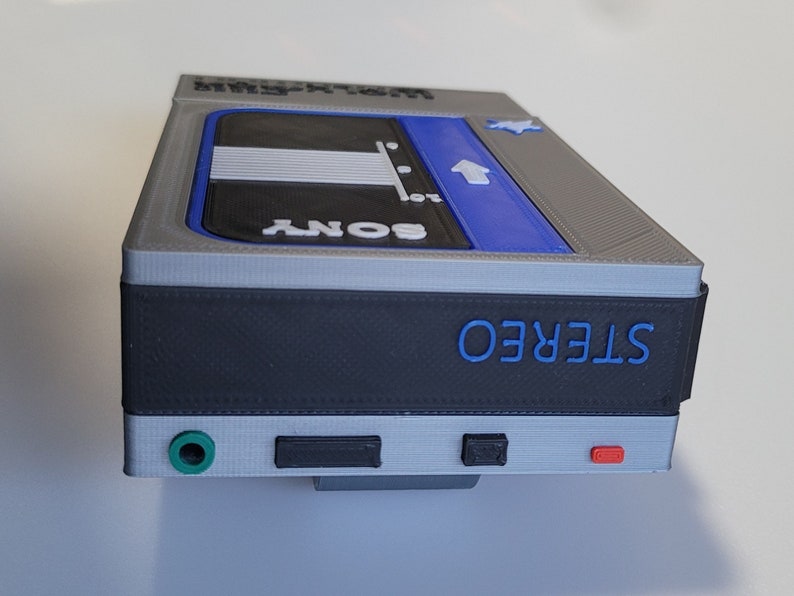 May include: A gray, black, and blue Sony Walkman stereo cassette player. The top has the text "WALKMAN" and a blue section with an arrow. The front has a green jack, a black button, and a red button. The word "STEREO" is in blue.