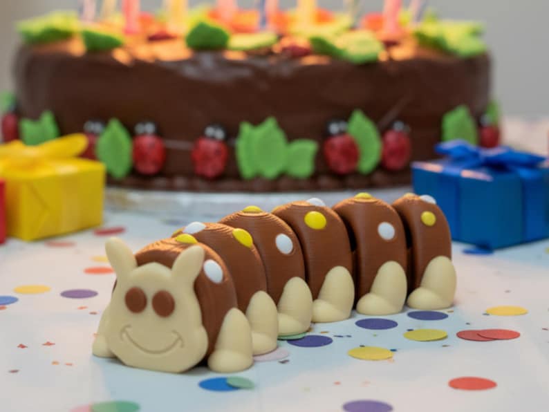 May include: A chocolate frosted birthday cake with candles, decorated with green leaves and red ladybugs. A brown caterpillar-shaped candy with white and yellow accents is in the foreground. Colorful confetti and wrapped gifts are also visible.