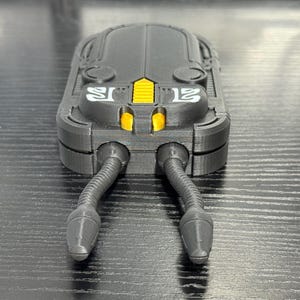 May include: A black, futuristic-looking device with a rectangular body and two flexible, tube-like appendages extending from the bottom. The device has yellow accents and the number "12" in white on the side.