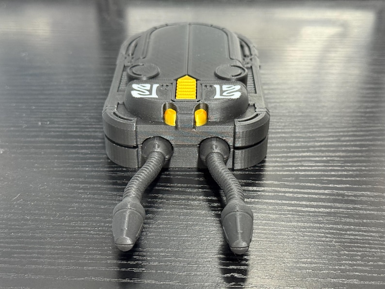 May include: A black, futuristic-looking device with a rectangular body and two flexible, tube-like appendages extending from the bottom. The device has yellow accents and the number "12" in white on the side.