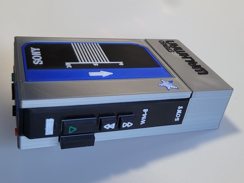 May include: A silver, black, and blue Sony Walkman WM-8, a vintage portable cassette player. The device features a blue accent, a graphic with white lines, and the Sony logo. The buttons are black, and the device is on a white surface.