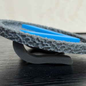 Puede incluir: Un objeto circular gris texturizado con una base curva, que sostiene dos piezas rectangulares azules. El objeto tiene una apariencia rugosa, similar a la piedra. La base es de color gris oscuro.
