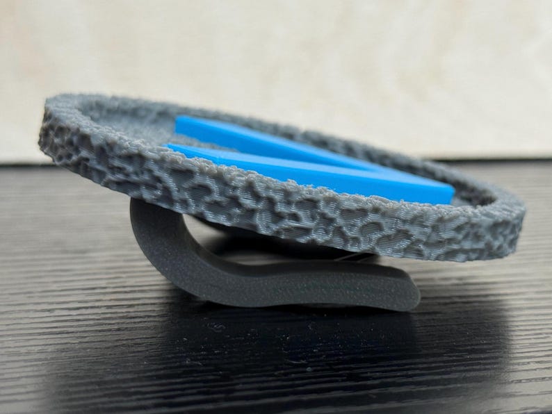Puede incluir: Un objeto circular gris texturizado con una base curva, que sostiene dos piezas rectangulares azules. El objeto tiene una apariencia rugosa, similar a la piedra. La base es de color gris oscuro.