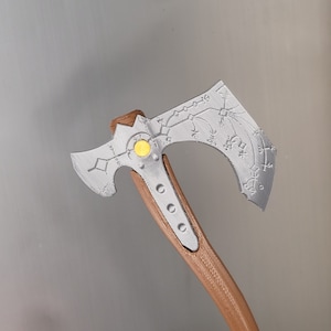 Op de afbeelding: Een zilveren bijl met een houten steel. De bijlkop heeft een decoratief ontwerp met een gele cirkel in het midden.