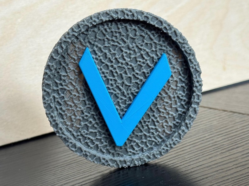 Puede incluir: Un objeto redondo gris impreso en 3D con una superficie texturizada y una forma de 'V' azul brillante en el centro. El objeto tiene un borde en relieve y con patrones. El dise&ntilde;o es simple y moderno.