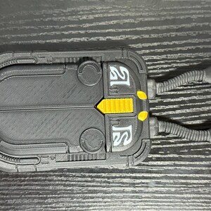 May include: A black and gray rectangular device with yellow accents and two flexible tubes extending from one side. The device has a textured surface and white text. The overall design suggests a futuristic or industrial aesthetic.
