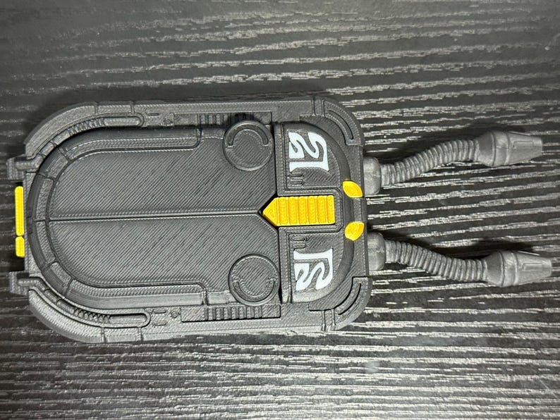 May include: A black and gray rectangular device with yellow accents and two flexible tubes extending from one side. The device has a textured surface and white text. The overall design suggests a futuristic or industrial aesthetic.