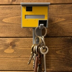 K&ouml;nnte beinhalten: Ein gelb-grauer Schl&uuml;sselhalter in Form eines Blitzers, an einer Holzwand befestigt. Der Schl&uuml;sselhalter hat ein rechteckiges Design mit einem Schlitz f&uuml;r Schl&uuml;ssel und einem Ring, der mehrere Schl&uuml;ssel h&auml;lt.
