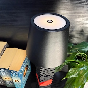 May include: A black table lamp with a white light on top. The lamp has a power button on the top. The lamp is sitting on a table with books and a plant.