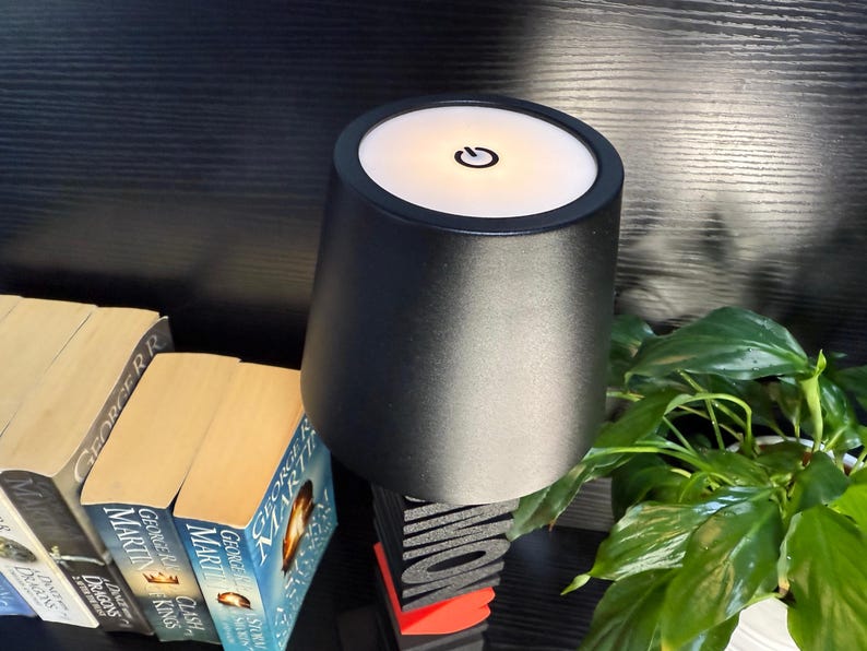 May include: A black table lamp with a white light on top. The lamp has a power button on the top. The lamp is sitting on a table with books and a plant.
