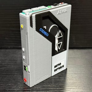 May include: A gray and black portable cassette player with the brand name "AIWA" on top. The player has a black front panel with the word "STEREO" in blue and "AUTO REVERSE" in white. It features buttons and ports in various colors.