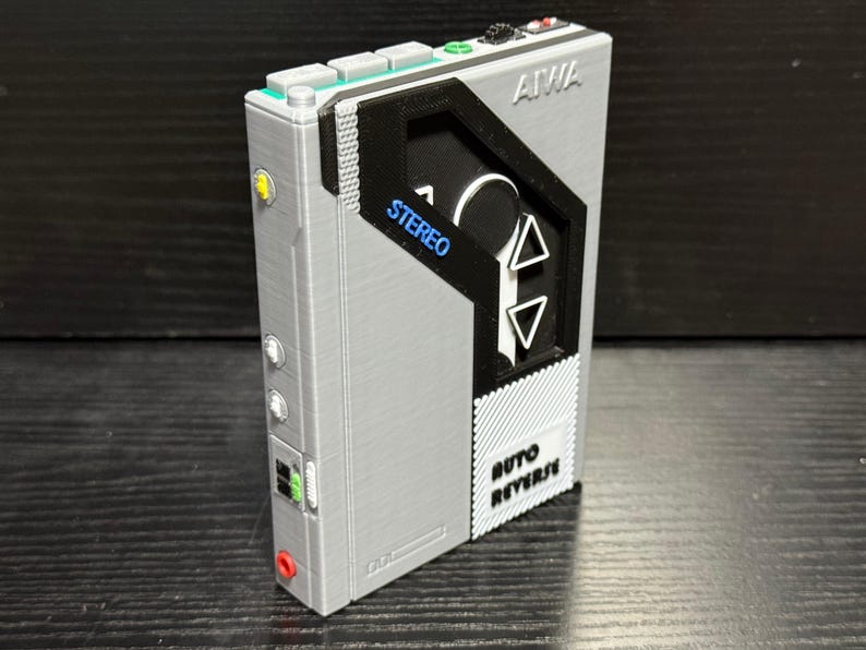 May include: A gray and black portable cassette player with the brand name "AIWA" on top. The player has a black front panel with the word "STEREO" in blue and "AUTO REVERSE" in white. It features buttons and ports in various colors.
