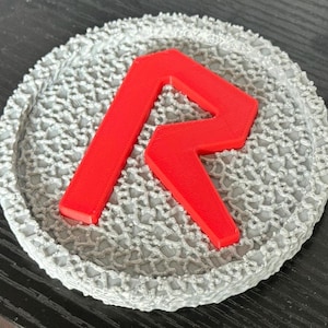 May include: A round, gray coaster with a textured, lace-like pattern. A bright red letter "R" is centered on the coaster. The coaster is likely made of a 3D-printed material.
