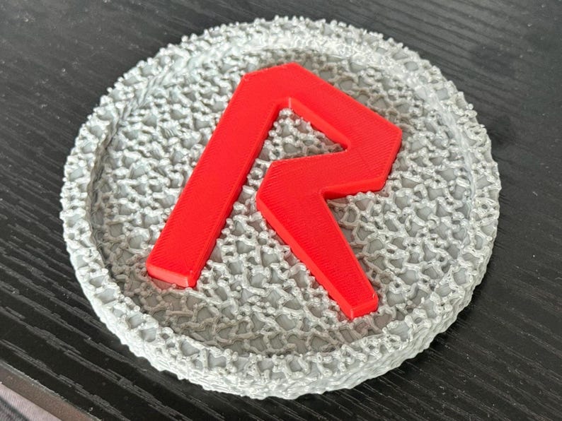 May include: A round, gray coaster with a textured, lace-like pattern. A bright red letter "R" is centered on the coaster. The coaster is likely made of a 3D-printed material.