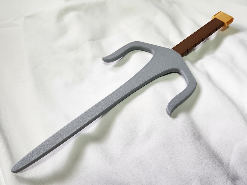 May include: A gray sai weapon with a brown handle and a gold-colored end. The sai has a three-pronged design and is set against a white background. The weapon appears to be made of plastic or a similar material.
