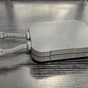 May include: A gray, rectangular object with two articulated arms extending from one side. The object has a textured surface and appears to be made of plastic or a similar material. The arms have a ribbed design.