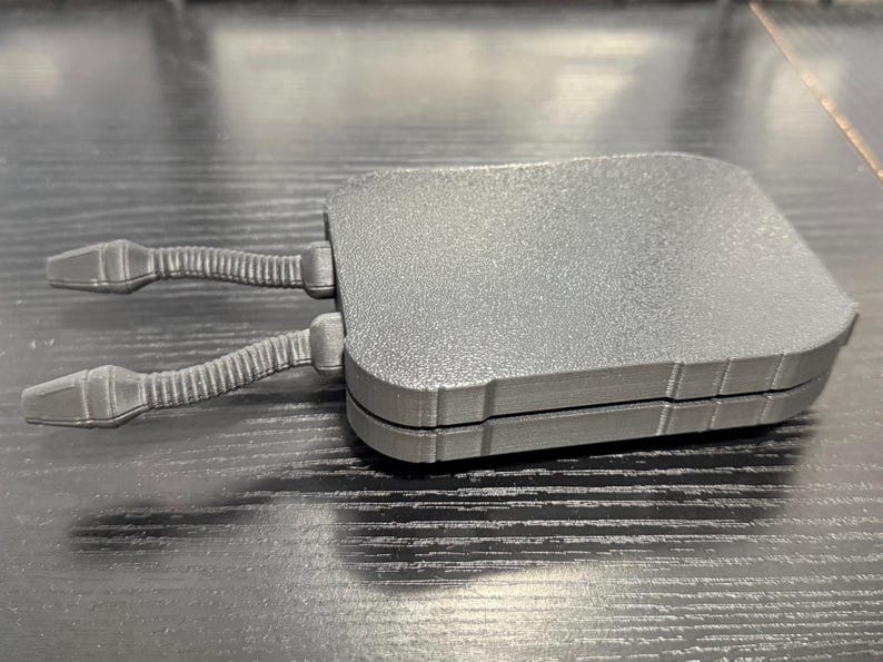 May include: A gray, rectangular object with two articulated arms extending from one side. The object has a textured surface and appears to be made of plastic or a similar material. The arms have a ribbed design.