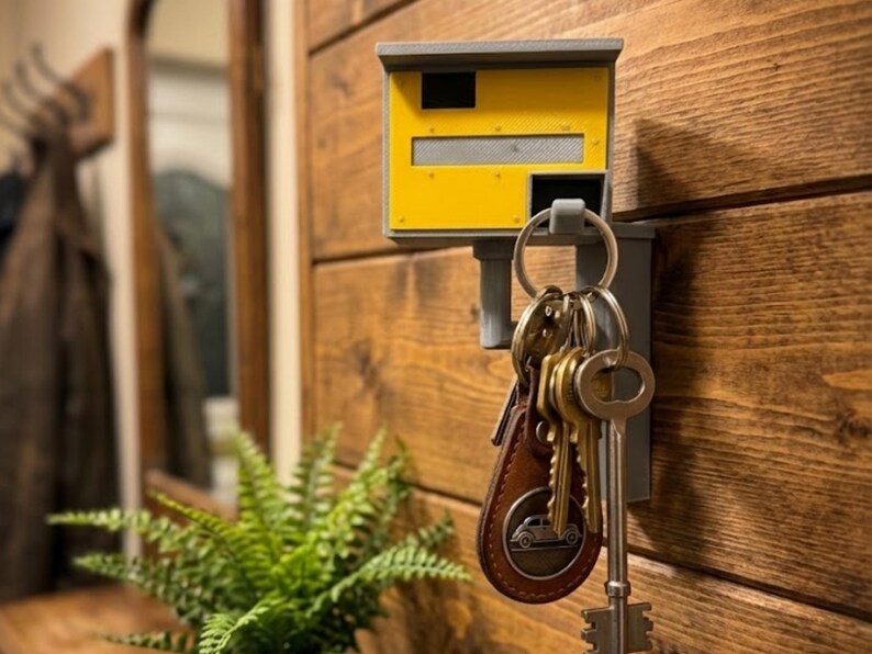 K&ouml;nnte beinhalten: Ein gelb-grauer Schl&uuml;sselhalter in Form eines Blitzers, montiert an einer Holzwand. Eine Sammlung von Schl&uuml;sseln h&auml;ngt an dem Halter, darunter ein Lederanh&auml;nger mit einem Autoemblem. Eine Farnpflanze befindet sich im Vordergrund.