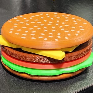 May include: A toy hamburger made of plastic with a brown bun, yellow cheese, red tomato, green lettuce, and a brown patty.