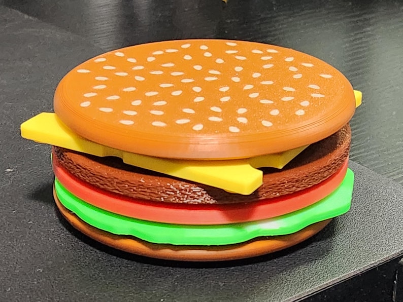 May include: A toy hamburger made of plastic with a brown bun, yellow cheese, red tomato, green lettuce, and a brown patty.