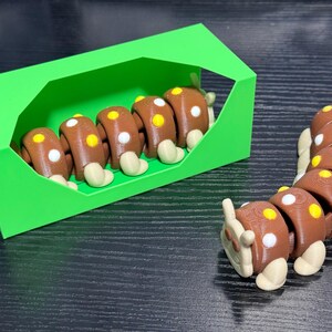 May include: A brown and white segmented caterpillar toy with yellow and white spots, and a green box. The caterpillar is made of several connected segments, with rounded feet. The toy is on a dark surface.