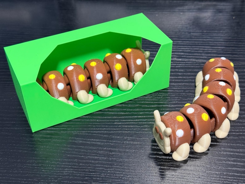 May include: A brown and white segmented caterpillar toy with yellow and white spots, and a green box. The caterpillar is made of several connected segments, with rounded feet. The toy is on a dark surface.