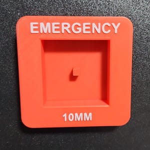 May include: Red plastic square with the word "EMERGENCY" in white lettering. There is a square cutout in the center with a small square in the middle. The bottom of the square has the text "10MM" in white lettering.