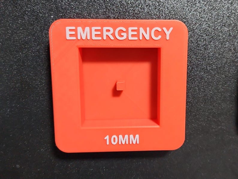 May include: Red plastic square with the word "EMERGENCY" in white lettering. There is a square cutout in the center with a small square in the middle. The bottom of the square has the text "10MM" in white lettering.