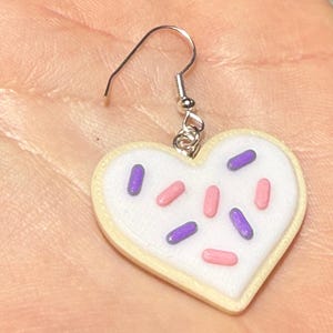 May include: A heart-shaped earring resembling a sugar cookie. The cookie has a white frosting base with pink and purple sprinkles. The earring is attached to a silver-colored fish hook.