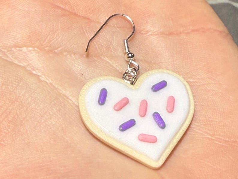 May include: A heart-shaped earring resembling a sugar cookie. The cookie has a white frosting base with pink and purple sprinkles. The earring is attached to a silver-colored fish hook.