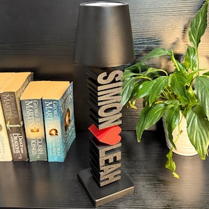 May include: A black table lamp with a black lampshade and a wooden base. The base is decorated with the names "SIMON" and "LEAH" in large, black letters. A red heart is placed between the names. The lamp is sitting on a black surface.