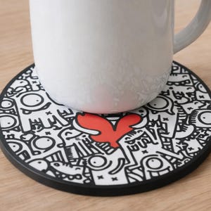 以下が含まれることがあります： 白いセラミックマグが丸いコースターの上に置かれています。コースターは、中央に赤いハートの形をした白黒の落書きデザインが特徴です。コースターには黒い縁があります。マグは明るい木製の表面にあります。