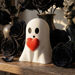 May include: A white ghost-shaped figurine with a red heart, set on a wooden surface. Black roses and vases are in the background, along with a gargoyle statue. The overall theme is gothic and romantic.