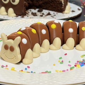 May include: A whimsical chocolate caterpillar cake on a white plate, decorated with colorful sprinkles. The caterpillar has brown segments with white and yellow accents, and a smiling face. A chocolate cake with white accents is in the background.