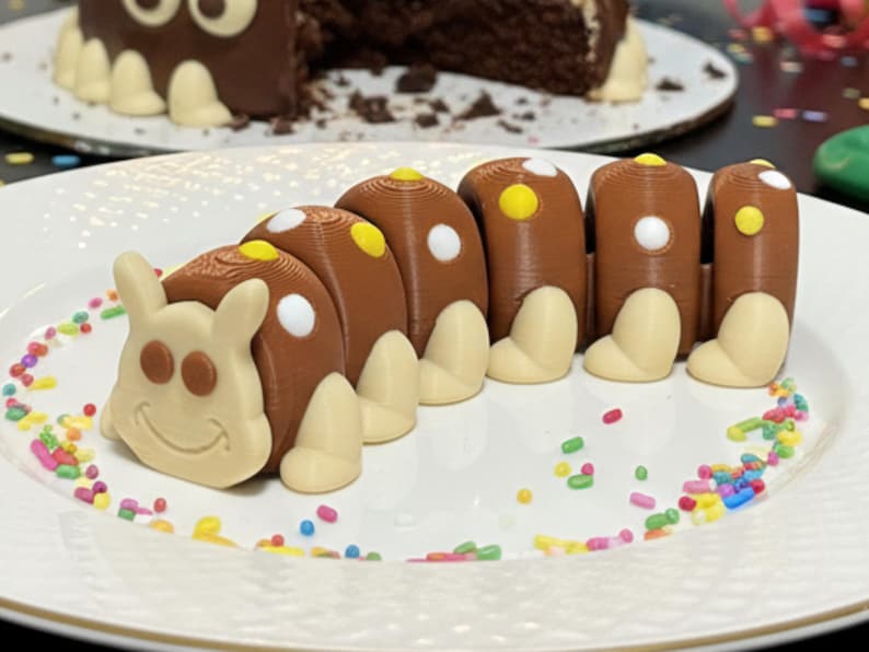 May include: A whimsical chocolate caterpillar cake on a white plate, decorated with colorful sprinkles. The caterpillar has brown segments with white and yellow accents, and a smiling face. A chocolate cake with white accents is in the background.