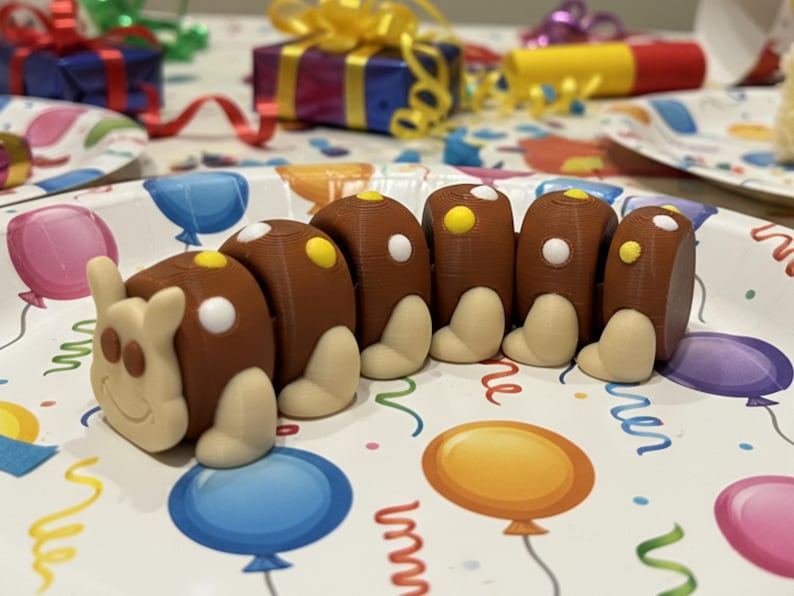 May include: A brown caterpillar-shaped dessert sits on a party plate decorated with balloons and streamers. The caterpillar has white and yellow accents. Other party decorations, including wrapped gifts, are in the background.