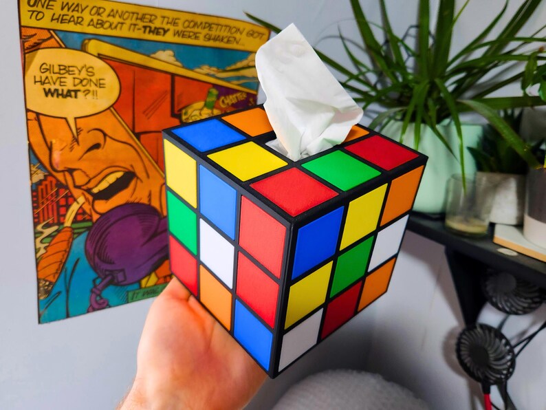 May include: A tissue box holder shaped like a Rubik's Cube with a white tissue box inside. The cube has the classic color scheme of red, blue, yellow, green, orange, and white.