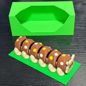 May include: A 3D-printed brown caterpillar toy with white and yellow spots, resting on a green rectangular base. A bright green box is in the background. The toy is designed with segmented body parts and rounded feet.