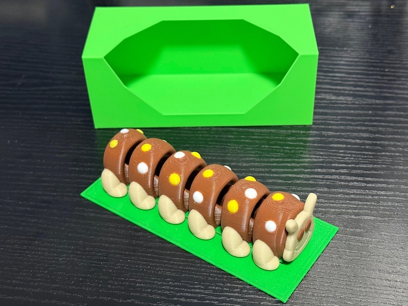 May include: A 3D-printed brown caterpillar toy with white and yellow spots, resting on a green rectangular base. A bright green box is in the background. The toy is designed with segmented body parts and rounded feet.