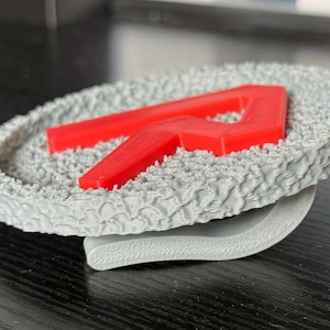 May include: A gray, textured coaster with a raised red letter "R" in the center. The coaster has a unique, patterned surface and a curved, gray base. The design is modern and minimalist.